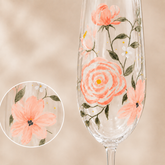 A clear stemmed glass with hand-painted coral pink flowers, green leaves, and small white blossoms, shown against a soft beige background with a circular close-up inset of one flower detail.