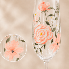 A clear stemmed glass with hand-painted coral pink flowers, green leaves, and small white blossoms, shown against a soft beige background with a circular close-up inset of one flower detail.
