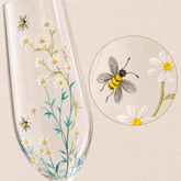 A clear stemmed glass decorated with painted white daisies, yellow centers, green stems, and two gray-and-yellow bees, shown on a warm beige background with a circular close-up inset of one bee and flower detail.