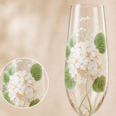 A clear champagne flute with hand-painted white flowers and green leaves, shown against a soft beige background with a circular close-up inset of the floral design.