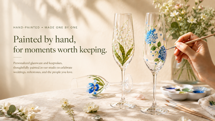 Two hand-painted champagne flutes on a table, one with white lily of the valley stems and the other with blue flowers and green leaves, with a hand painting the second glass. Text on the left reads 'Hand-Painted • Made One by One' and 'Painted by hand, for moments worth keeping.