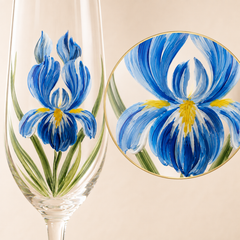 A clear stemmed glass with a hand-painted blue iris flower and green leaves, shown alongside a circular close-up inset of the floral artwork on a beige background.