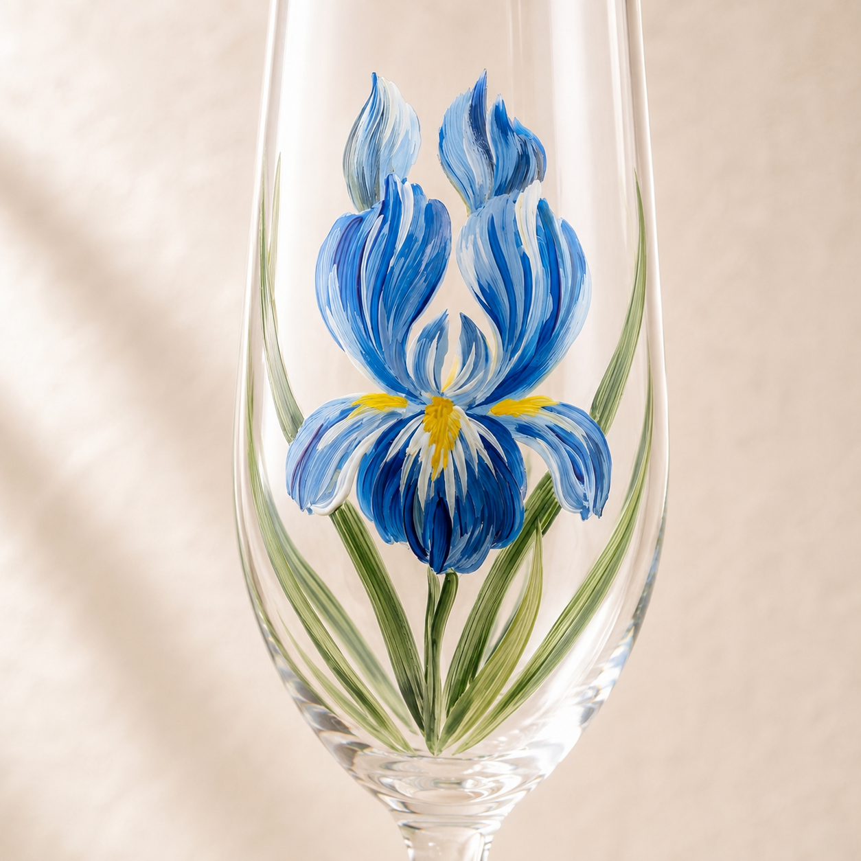 Close-up of a hand-painted blue iris flower with green leaves on a clear champagne flute
