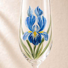 Close-up of a hand-painted blue iris flower with green leaves on a clear champagne flute
