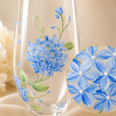 A clear glass vase with hand-painted blue hydrangea flowers and green leaves, shown on a soft beige fabric background with a circular close-up inset of the floral design.