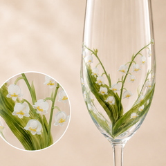 A clear stemmed glass with hand-painted lily of the valley flowers and green stems, shown against a soft beige background with a circular close-up inset of the floral design.