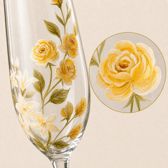 A clear glass wine flute decorated with hand-painted yellow roses, green leaves, and small white flowers, shown on a warm beige background with a circular close-up inset of the floral design.