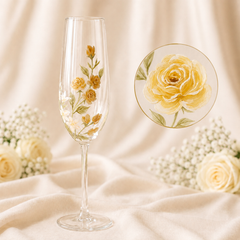 A clear champagne flute with delicate yellow and green floral hand-painted detailing, displayed on draped cream fabric alongside white roses and a circular inset showing a close-up of the floral design.