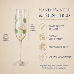 A hand-painted and kiln-fired champagne flute with white floral and green leaf designs, shown on a beige background with text detailing its dimensions and features.