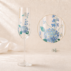 A clear stemmed glass with a floral print of blue forget-me-nots and green leaves, shown on a warm beige textured background with a circular close-up inset of the decoration.